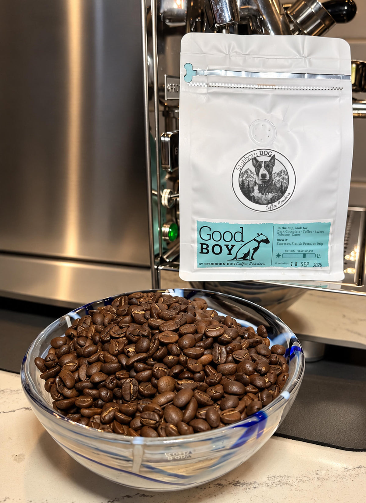 A bag of Good Boy Medium-Dark Roast coffee beside a glass bowl of roasted beans, in front of an espresso machine
