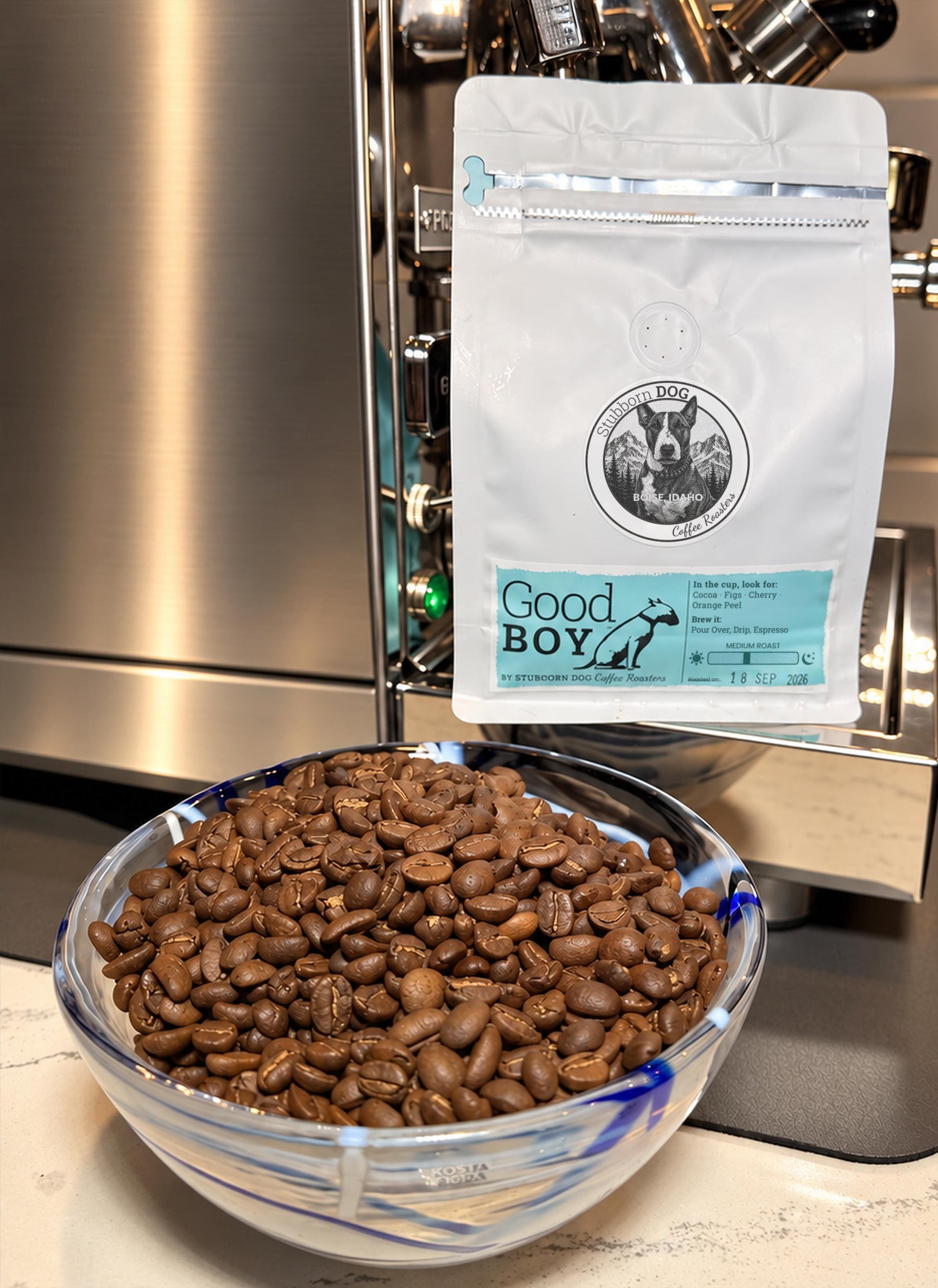 A bag of Good Boy Medium Roast coffee beside a glass bowl of roasted beans, in front of a stainless-steel espresso machine